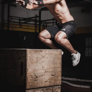 Shirtless man showcasing strength and fitness with a box jump in an indoor gym setting.