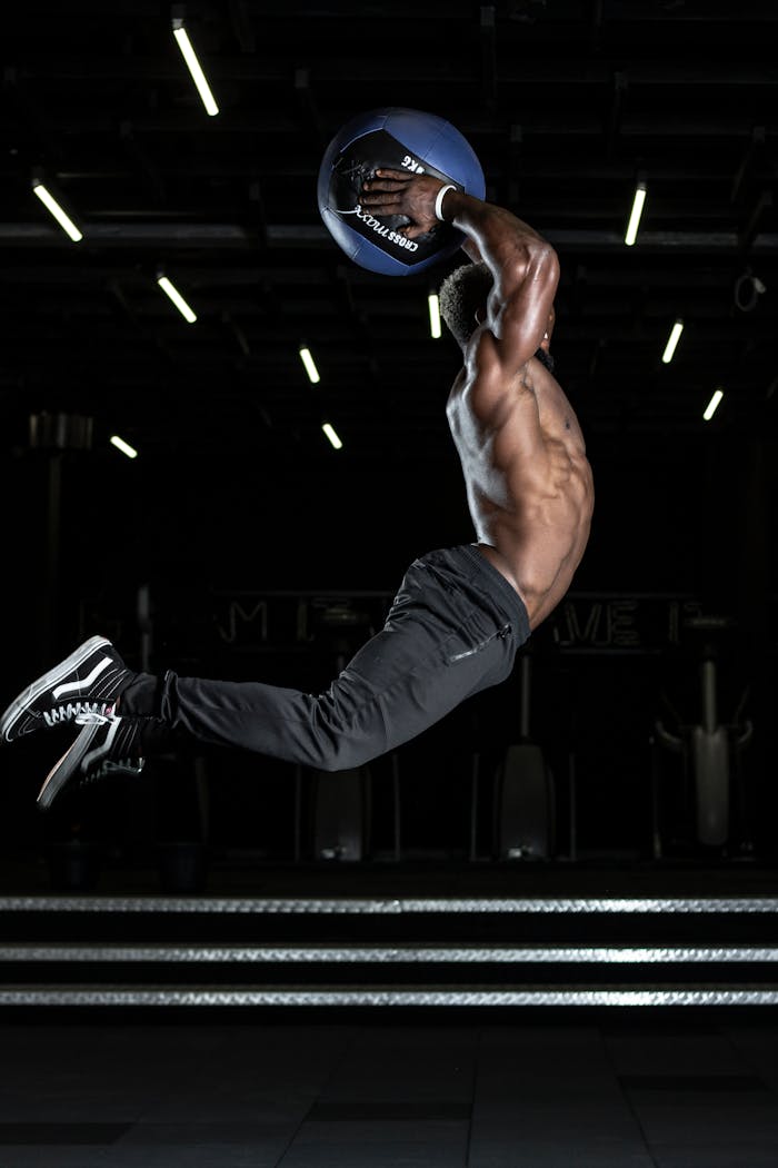 Muscular athlete performing a high jump with a medicine ball in a gym, showcasing strength and fitness.